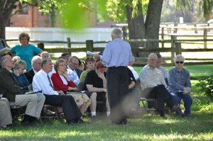 A photo of a group of people sitting outside listening to a person speaking.